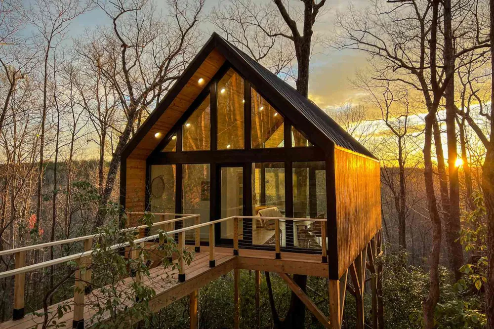 A modern A-frame glass cabin elevated on stilts among bare trees, glowing warmly at sunset