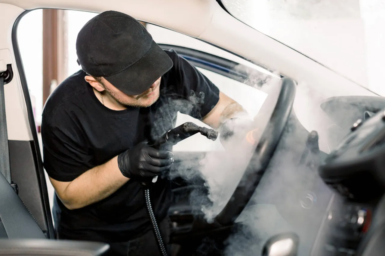 An auto detailer in gloves using a steam cleaner to deep clean a car's interior through the open door
