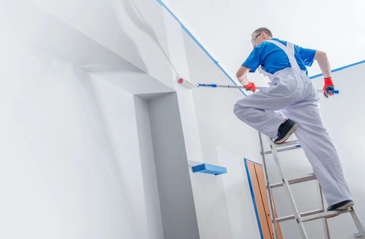 A painter on a ladder rolling a fresh coat of paint onto an interior wall lined with painter's tape