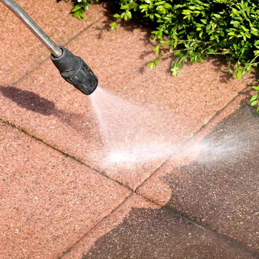 Close-up of a pressure washer nozzle blasting dirt from red patio pavers beside a green hedge