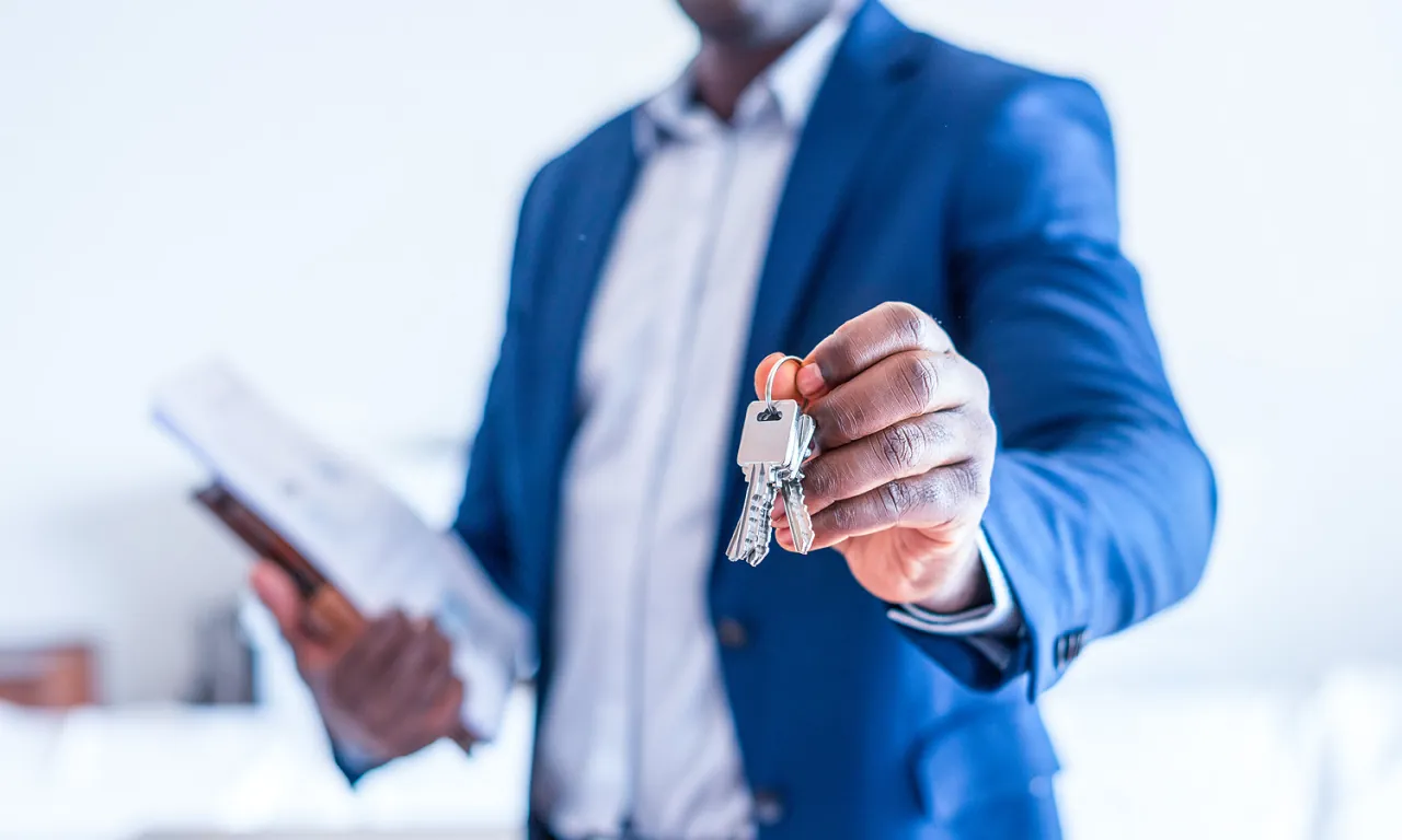 A real estate agent in a blue suit holding out a set of house keys to a new homeowner