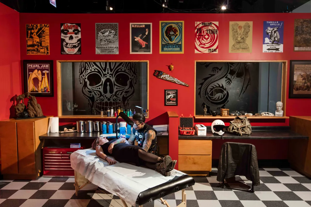 Interior of a tattoo studio with red walls, framed posters, and an artist tattooing a reclining client