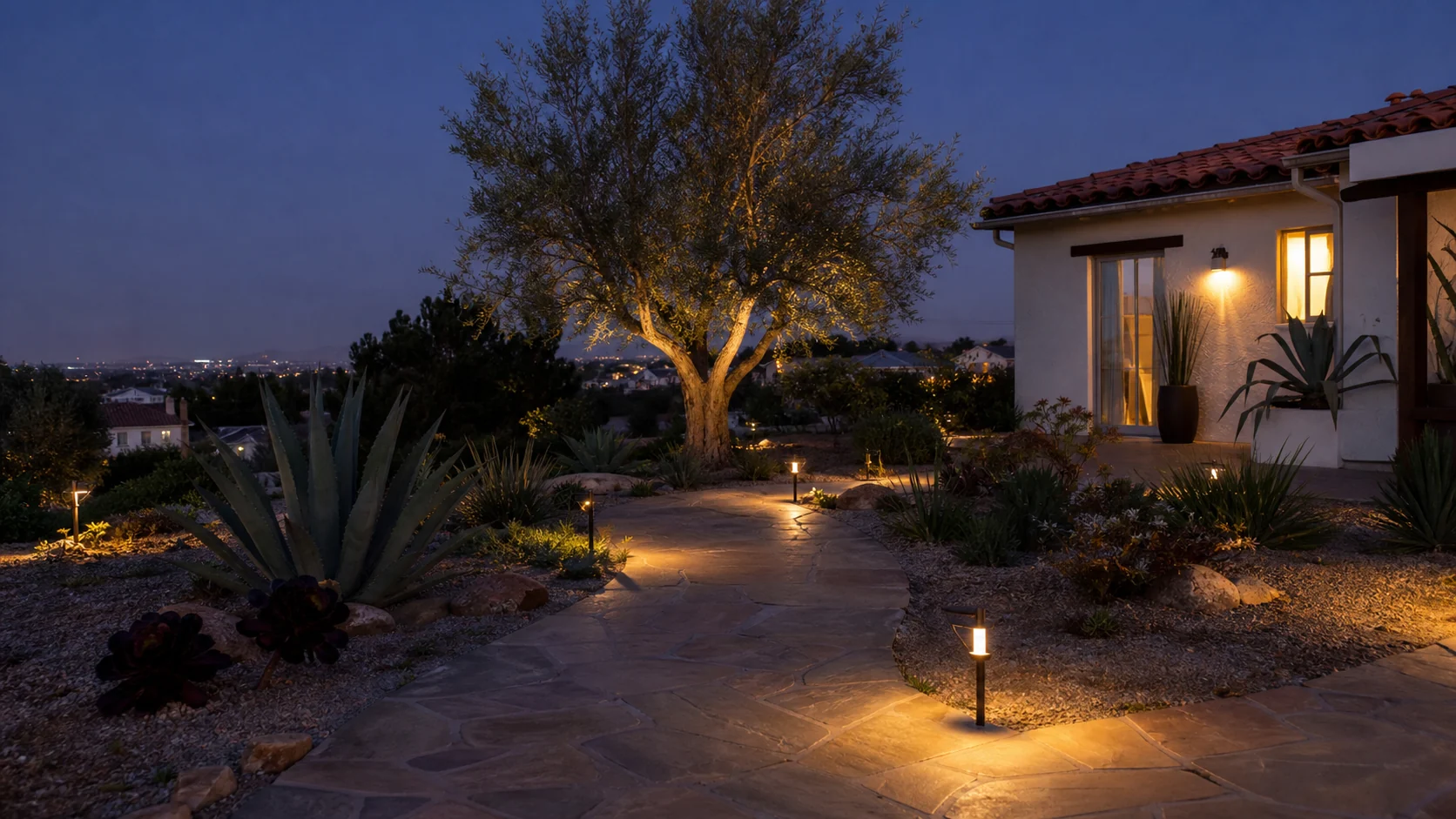 A Santa Ana front yard at dusk with warm low-voltage path lighting tracing a flagstone walkway past agave and an olive tree, with a stucco home on the right